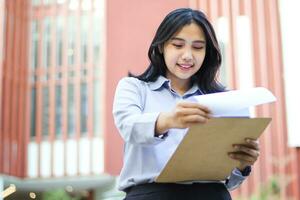 sonriente asiático mujer de negocios gerente leyendo reporte papel mientras caminando en al aire libre, contento vendedora supervisor muy ventas datos mientras caminando terminado ciudad edificio foto