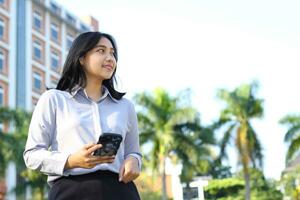 smiling busy asian business woman walking on city streets using application on smartphone wearing formal suit standing over urban building with green plant background looking away photo