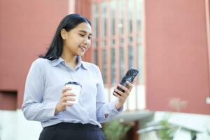 confident asian business woman walking over outside office building using smartphone and drink coffe with smile, beautiful executive manager standing in outdoors surfing social media photo