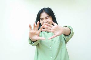 carefree asian woman raising palms forward showing stop gesture wearing green oversized shirt looking at camera with laughing excitement face expression isolated on white background photo