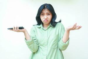 portrait of confused asian woman wearing green shirt shrug shoulders and spreads palms holding smartphone looking at camera worried standing over isolated background, considering gesture photo