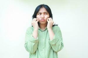 upset asian woman with dark hair touch her cheek wit curled up figer and looking away dissatisfied wearing green oversized shirt isolated on white background photo