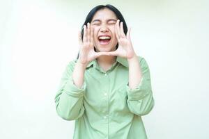 confident asian woman with dark hair raising palms over her open mouth and shouting announce product with eye closed wearing green oversized shirt isolated on white background photo