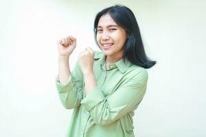 playful success dark hair asian woman laughing and dancing to celebrate champion win victory by raise fist wearing green oversize shirt standing over isolated white background, looking at camera photo