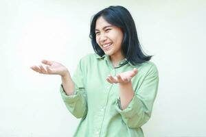 carefree delighted dark hair asian woman raising palms and looking away surprised wearing green oversized shirt isolated on white background, receiving gift gesture photo