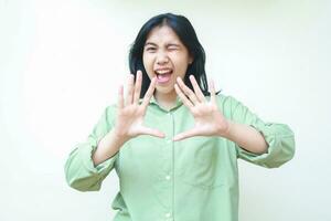 happy asian woman surprised looking at camera with raising palms show stop gesture, warning, flirting and blink eye, wearing green oversized shirt isolated on white background photo