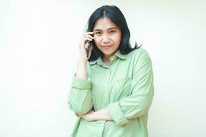 happy calm asian woman wearing green oversize shirt talking on smartphone with folding arms looking at camera with serious face isoalted on white background photo
