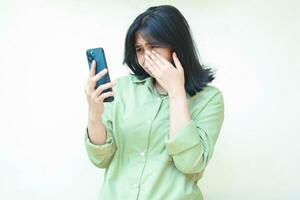 sad asian woman wipes her tears after cry for using social media on smartphone hold on hands wearing green oversized shirt standing over isolated background. Mental health concept, bullying photo