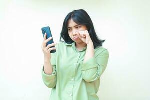 sad asian woman wipes her tears after cry for using social media on smartphone hold on hands wearing green oversized shirt standing over isolated background. Mental health concept, bullying photo