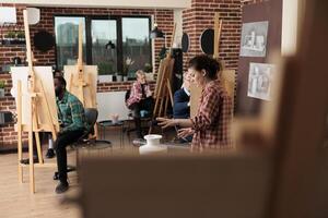 Group of different aged people sitting at easels studying drawing at group art class. Young woman teacher explaining still life sketching techniques to students. Social activities for adults photo