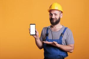 Construction worker holding cell phone with empty screen for advertising in front of camera. Professional builder with hard hat against yellow background in studio shot. photo