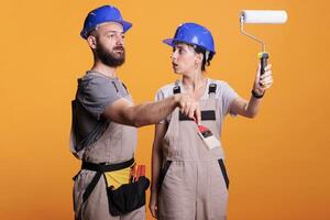 Young construction workers posing with painting tools, standing over yellow background. Team of constructors holding paintbrush and roller brush to work on refurbishment project in studio. photo