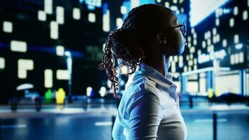 Smiling african american woman wandering around city boulevards during nighttime, enjoying life. Businesswoman strolling around in office buildings urban center, surrounded by bokeh street lights photo