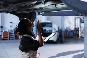 africano americano mecánico usos Bosquejo tableta mientras cambiando antiguo coche freno fluido con nuevo uno en reparar tienda. bipoc mujer participación aislado pantalla dispositivo en garaje mientras haciendo vehículo chequeo foto