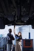 Engineer in car service uses tablet to calculate invoice after fixing broken automobile tires. Worker uses device to inform client of final costs after mending vehicle suspended on overhead lift photo