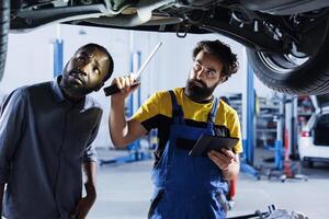 Repairman in car service uses tablet to calculate invoice after fixing broken automobile wheels. Professional uses device to inform client of final costs after mending vehicle suspended on overhead photo