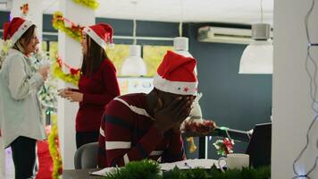 African american employee in Christmas decorated office working, trying to concentrate while carefree coworkers enjoy coffee break together. Worker with santa hat frustrated by distracting colleagues photo