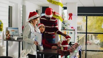 Staff gathered together at desk brainstorming ideas for project in Christmas decorated office, zoom out. Multiethnic group of workers in workplace adorn with xmas tree and garlands photo