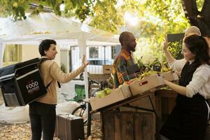 Young marketplace stand vendor giving food order to delivery worker, working on delivering fresh produce from farm. Female farmer talking to courier on bike, deliver healthy bio products. photo