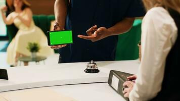 Guest showing phone with greenscreen, holding mobile phone template with isolated chromakey display to hotel staff in lobby. Man traveller using mockup copyspace at front desk. photo