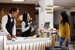 asiático personal haciendo un reserva para cliente a el frente escritorio. sonriente conserje comprobación en un cliente. mujer turista recepción Consejo desde simpático personal. turista siendo bienvenido a elegante hotel. foto