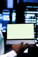 Expert examining server cabinets performance trends using green screen device. Serviceman uses chroma key laptop to pinpoint data center operational issues arising due to insufficient memory, close up photo