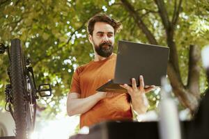 Healthy dedicated caucasian man searching on laptop to fix broken modern bicycle. Active young male cyclist ensures bike components are secure for summer outdoor leisure cycling. photo