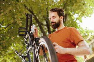 activo y sano masculino ciclista inspeccionando su bicicletas rueda para daños y perjuicios en el patio trasero. imagen retrata joven caucásico hombre utilizando lubricante a reparar roto componente asegurando un seguro y seguro paseo foto