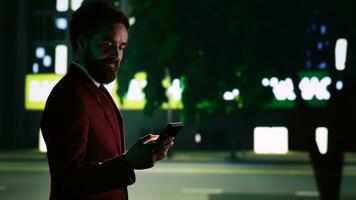 Classy businessman checks messages on night walk, strolling around downtown district and looking for directions on sidewalk. Confident man walking around city, evening promenade. Handheld shot. photo