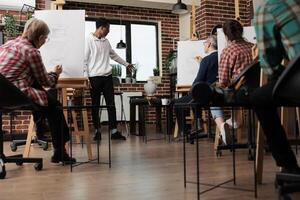 Group of people drawing with pencils on canvas, attending art class. African American guy teacher standing in classroom showing still life object, explaining how to draw shapes and proportions photo