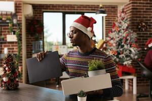 trastornado empresa trabajador embalaje pertenencias y ordenador portátil después siendo despedido en Navidad víspera. infeliz africano americano hombre experimentando desempleo después consiguiendo despedido durante invierno fiesta foto