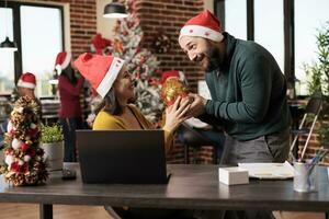 Excited woman employee laughing while receiving christmas gift bag from colleague in santa hat. Cheerful caucasian man greeting coworker with xmas and giving present in coworking space photo