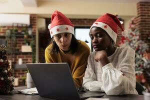 Two women coworkers checking project report on laptop and celebrating xmas in decorated office. African american and caucasian employees in santa hats working on startup task together photo