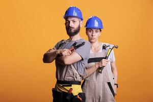 Confident professional renovators posing with pair of pliers and hammer, holding slegdehammer and renovating tools. Man and woman working as constructors with overalls and helmet. photo