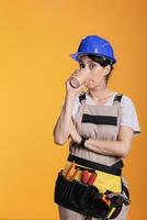 Female construction worker enjoying hot coffee during break time at work. Professional woman renovator wearing belt with tools and protective helmet in studio shot against yellow background. photo