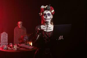 Beautiful woman working on laptop in studio, checking internet website online on wireless pc. Santa muerte model wearing halloween costume and dios de los muertos skull make up. photo