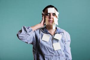 Portrait of stressed depressed executive manager overwhelmed of work with sticky notes on her face, standing over isolated background in studio. woman using memory papers to write down job details photo