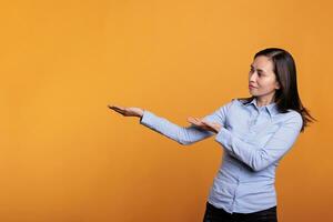 Asian woman pointing at presentation icon on camera, doing advertisement and showing promotional product. Model posing over yellow background in studio showing confident girl doing ad. photo