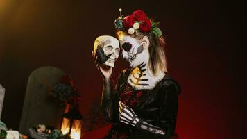 Glamorous woman acting creepy with skull in hand, holding black roses and wearing scary make up. Looking like santa muerte with body art on holy mexican celebration, being flirty. Handheld shot. photo