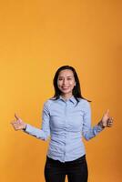 Smiling asian woman doing thumbs up gesture in front of camera showing approval and agreement gesture with fingers in studio. Satisfied model gesturing okay and positive symbol over yellow background photo
