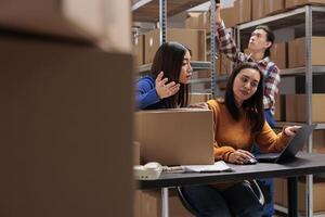 Storehouse employees checking customer delivery satisfaction on laptop and arguing about negative feedback. Postal service asian women coworkers managing shipment from warehouse computer photo