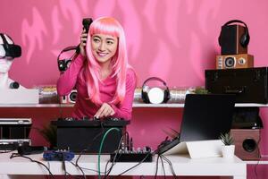 Artist with pink hair mixing techno song at professional turntables listening music with headphone while doing performance in club during nighttime. Woman recording album using audio equipment photo