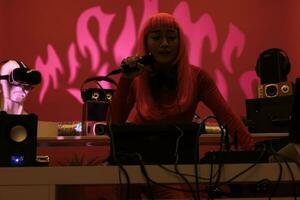 alegre artista con rosado pelo en pie a DJ mesa jugando electrónico música con tocadiscos, disfrutando a realizar a noche en club. asiático ejecutante teniendo divertido con aficionados durante techno concierto foto