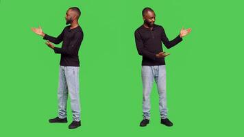 African american man creating advertisement in studio, pointing to left or right sides for ad presentation. Young male model in jeans and shirt presenting something on full body greenscreen. photo