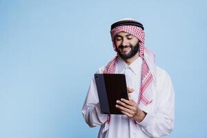 Smiling man wearing muslim headdress and thobe holding digital tablet and tapping on touchscreen. Arab person scrolling internet page on portable gadget with cheerful expression photo