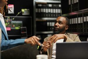 African american detective chatting with colleague while studying case reports. African american man and woman investigators cooperating and searching information about suspect together photo