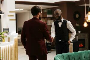 Diverse men shaking hands at hotel, african american concierge welcoming guest and offering his services. Bellhop provides excellent amenities to enhance business trip experience. photo