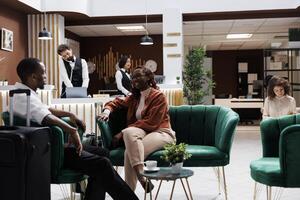 Two partners waiting to register at hotel front desk, sitting in lounge area spacious lobby. Man and woman relaxing on modern sofa before doing check in procedure at reception counter. photo