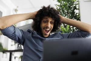 Arab start up company entrepreneur shocked with high sales growth while checking results on laptop. Executive manager celebrating effective marketing strategy in business office photo