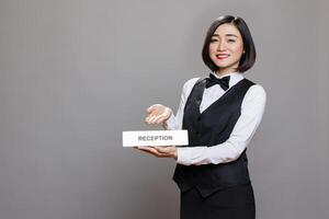 Hotel smiling woman employee showing reception plate studio portrait. Friendly asian receptionist wearing professional uniform pointing at steel name signage and looking at camera photo